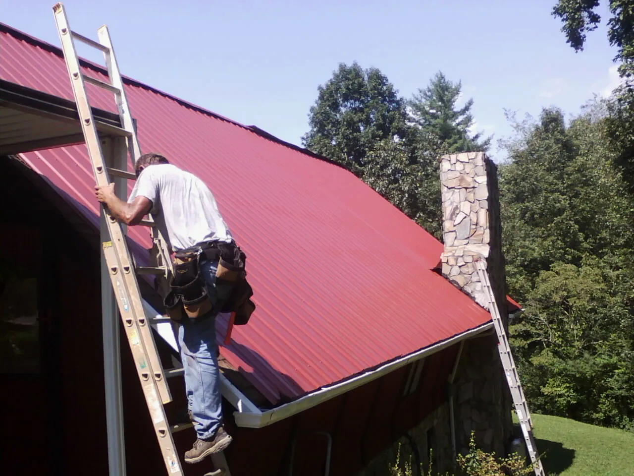 Roofer on a ladder beside a red metal roof and stone chimney — Soffit & Fascia Repair by Roofing Brothers in Reedville